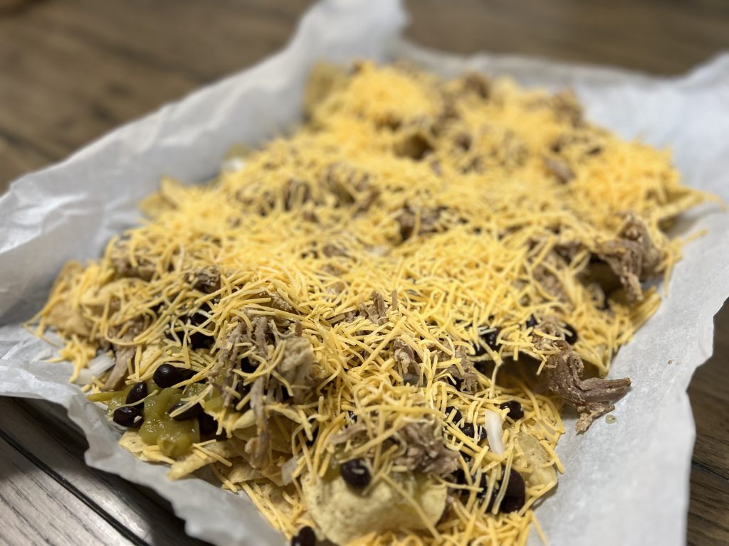 A picture of unbaked tortilla chips topped with cheese pulled pork, black beans, onions and green chiles on top of a baking sheet lined with parchment paper. 