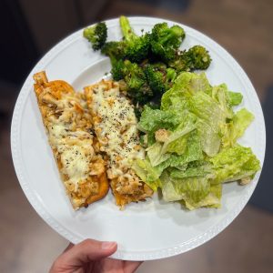 Picture of a hand holding a white plate that has the baked buffalo chicken flat bread pizza alongside a caesar salad (white dressing and green romaine lettuce) and pan fried broccoli with onions and parmesan cheese.